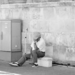 A black-and-white photo of an elderly person sitting on a small bench against a stone wall. They are wearing a hat, shirt, and slacks, and they are resting their head on their hand. A walking cane is leaning against a utility box beside them.