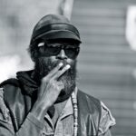 A man with a beard and sunglasses is smoking a cigarette. He is wearing a hat, a scarf, and a jacket. The photo is in black and white, and the background is blurred.