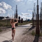 A person in a pink top and black bottoms holds a surfboard overhead, walking down a pathway bordered by metal scaffolding structures with a cloudy sky and industrial buildings in the background. The scene suggests a mix of nature and industrialization.