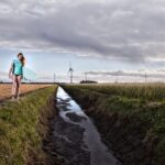 A person in a blue shirt walks along a narrow pathway flanked by fields near a water-filled ditch. They are holding a surfboard and looking down. In the background, wind turbines stand tall under a cloudy sky.