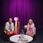 Two people sit on chairs near a round white table with magazines and vases. The man wears a beige blazer and red pants; the woman wears a pink top and patterned skirt. Behind them is a purple-lit curtain with a wooden stool, a lamp, and a floral arrangement on stage.