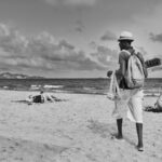 A person in a hat and shorts walks along a sandy beach carrying a tray with merchandise over their left shoulder. Other beachgoers are lounging on the sand or by the water under a cloudy sky.