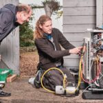 Two men working on an outdoor HVAC unit next to a brick wall and wooden shed. One man, standing, observes while the other, kneeling, uses a tool on the open unit. Various tools and equipment are scattered around them. The environment appears to be residential.