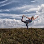 A woman performs a standing yoga pose on top of a grassy hill under a partly cloudy sky. She balances on one leg while holding the other leg behind her with one hand and extending the opposite arm forward. The background features a scenic landscape with distant hills.