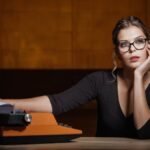 A woman with glasses and wearing a black top sits at a wooden table, resting her chin on her left hand, looking contemplative. Her right hand holds a piece of paper that is coming out of an orange typewriter. The background is warmly lit in a dark amber tone.