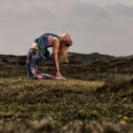 A woman in workout attire performs a camel yoga pose, leaning back with her hands on her heels and her head tilted back. She is practicing outdoors on a grassy hill under an overcast sky, with rolling hills in the background.