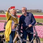 An older woman and man stand beside their bicycles in front of colorful tulip fields. The woman, dressed in a yellow coat and striped scarf, takes a selfie with a red smartphone, while the man, wearing glasses and a blue jacket, smiles beside her.