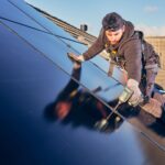 A worker, wearing a cap and safety harness, is kneeling on a tiled roof while installing a large solar panel. The worker is using a power drill to secure the panel, with clear blue skies in the background.