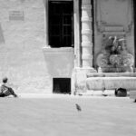 A person sits on the ground near an ornate stone fountain with statues, while a pigeon walks toward them in a spacious courtyard. The sign on the wall reads "MUSARAM SAN GORG." The scene is in black and white.