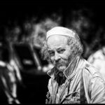 Black and white photo of an older man with curly hair and a mustache, wearing a cap and a jacket. He stands outdoors, looking slightly away from the camera with a thoughtful expression. Background is blurred, indicating possible urban or park setting.