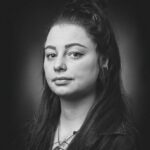 A black and white portrait of a person with long hair in a half-up bun. They have a neutral expression, a nose ring, and are wearing earrings. Their outfit includes a jacket, and a necklace with layered pendants is visible.