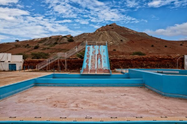 An abandoned water park features an empty pool with cracked surfaces in the foreground and a rusted water slide leading to the pool. Behind the park are rugged, eroded hills under a partly cloudy sky, creating a desolate and neglected atmosphere.