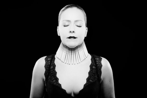 Black and white image of a person with eyes closed, wearing a detailed statement collar made of tubular elements. They are dressed in a lace-trimmed top, standing against a black background. The composition emphasizes simplicity and high contrast.