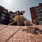 A person skateboards in mid-air between two metal railings on a brick slope in an urban area with large buildings and trees. The sky is clear and sunny, casting shadows on the ground. A bystander can be seen capturing the moment with a camera.