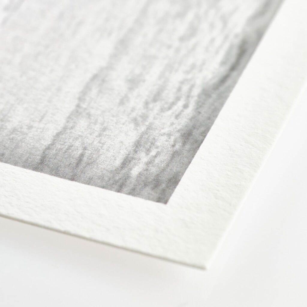 Close-up of the corner of a white-bordered photograph featuring a textured surface with varying shades of gray and subtle linear patterns, resembling a monochrome wood grain. The thick, textured print paper border contrasts with the smooth photo, adding an elegant touch to the overall presentation.