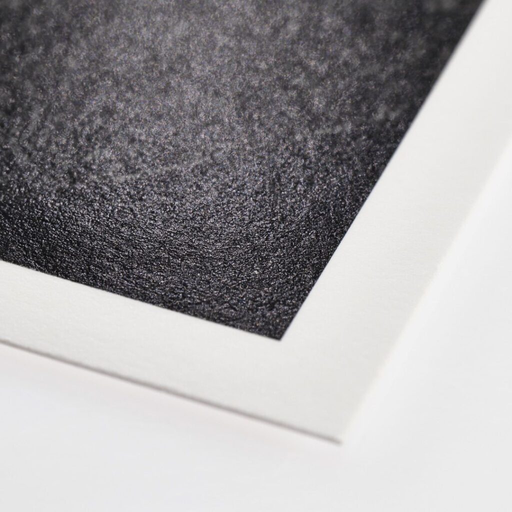 Close-up of a corner of textured black and white print papers. The black area is speckled and grainy, while the white border is smooth and clean. The angle of the image highlights the contrast between the two surfaces.