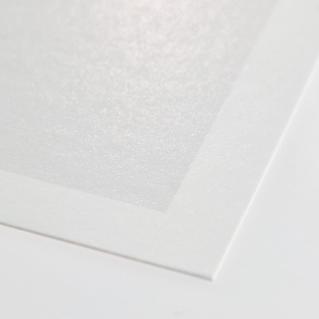 A close-up shot of the corner of a piece of white print paper on a white surface. The paper has a slightly textured finish, and the light reflects off the surface, giving it a subtle sheen.