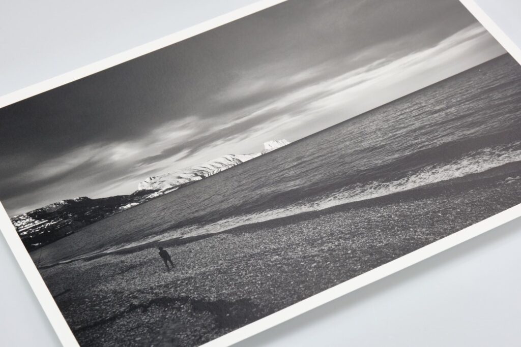 A black and white photograph, printed on high-quality print papers, depicts a lone individual standing on a pebbled beach, facing calm sea waves. Snow-covered mountains are visible in the background under a cloudy sky. The photo is slightly tilted.