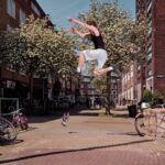 A person captured mid-air while performing a parkour jump between buildings in an urban setting. Another person on a low wall enthusiastically watches the jump. Bicycles are parked along the sidewalk, and there are flowering trees lining the street.