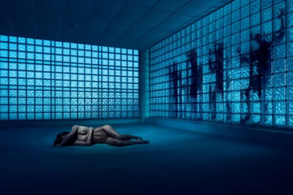 A person lies curled up on the floor in a dimly lit room with walls made of blue-tinted glass blocks. Shadows from the glass create a moody and contemplative atmosphere. The overall scene is somber and introspective.