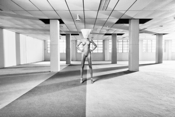 A black and white image features a person standing in an empty, open space with a tiled ceiling and numerous supporting columns. The individual is wearing high heels and a headpiece resembling a large cone or funnel, posing with hands on hips.