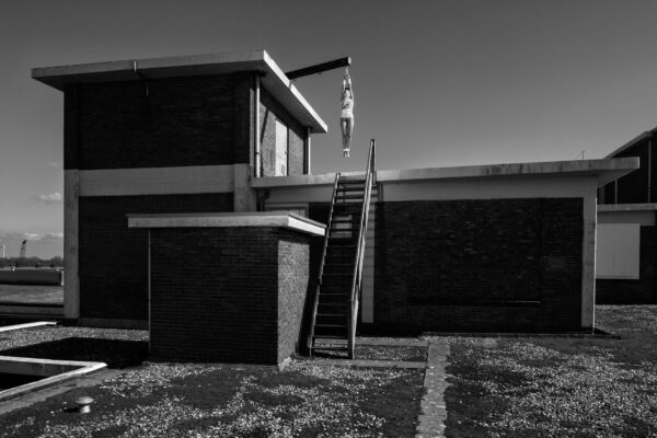 A black-and-white photo depicts a person hanging upside down from a metal beam attached to the roof of a two-story brick building, next to an industrial staircase. The surrounding area appears to be a deserted industrial site.