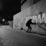 A person in a hoodie is spray painting graffiti on a concrete wall at night under streetlights. The graffiti spells out "ALMAAR" in bold, stylized letters. The scene is illuminated by a few streetlights along the road.