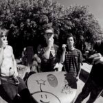 Four young men are gathered outdoors, standing around a graffitied trash can with a peace sign and smiley face. One is shirtless, two others have their hands near their faces, and the fourth is looking at a device in his hands. There are trees in the background.